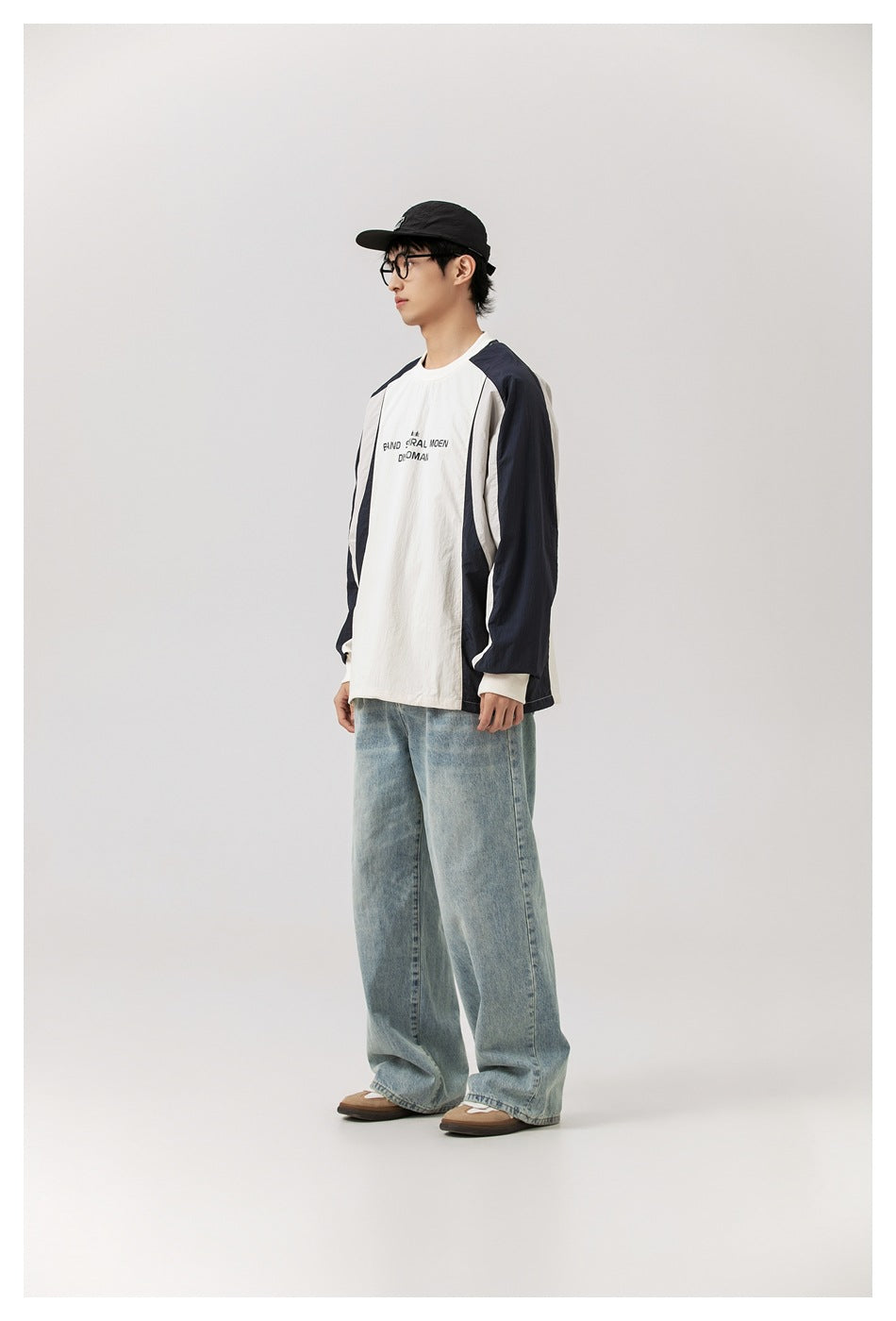 Model in color-block crew-neck sweatshirt and light-wash wide-leg jeans, side view, with black cap and glasses