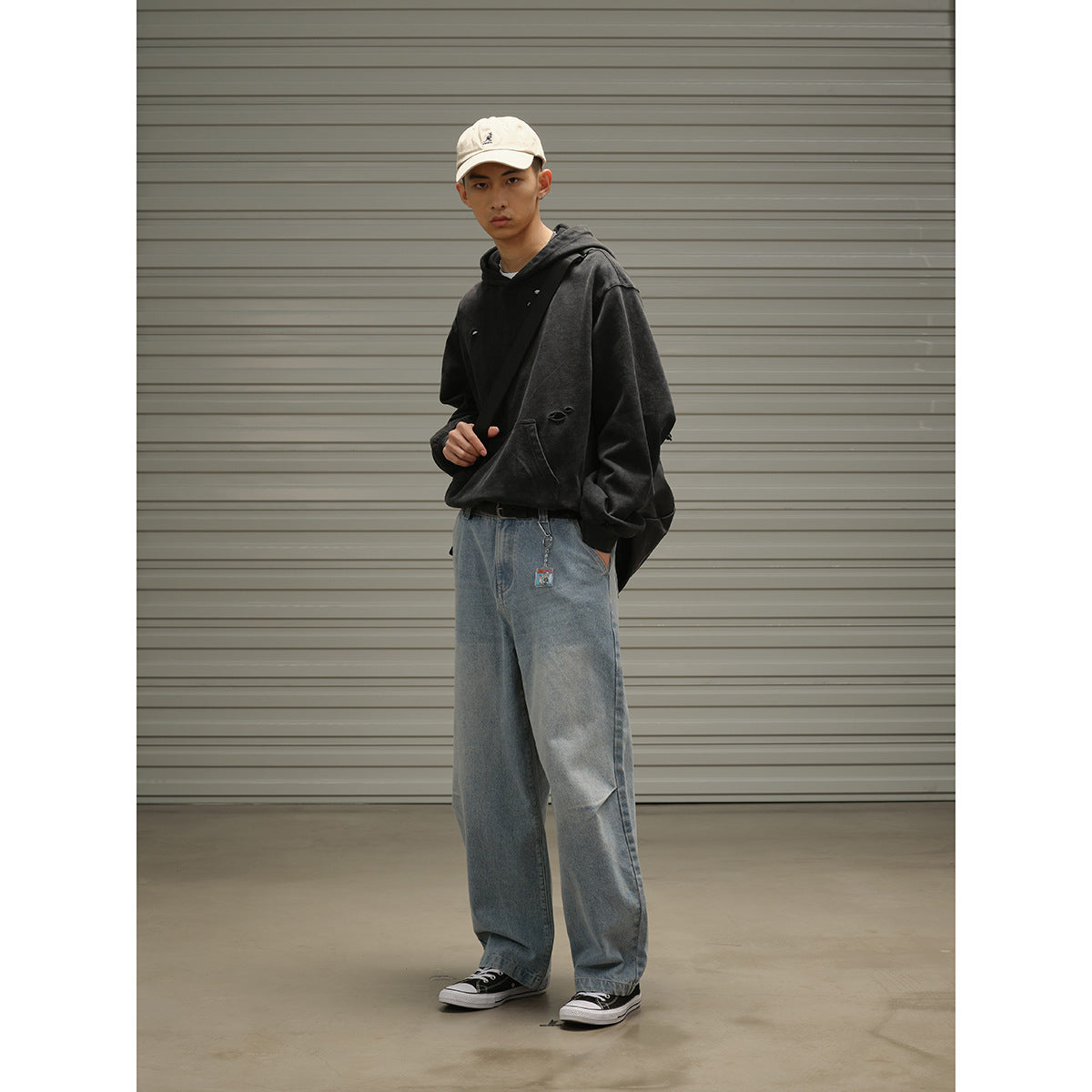 Model in a black distressed hoodie, loose blue jeans and cream cap, standing at an angle with a black crossbody bag.