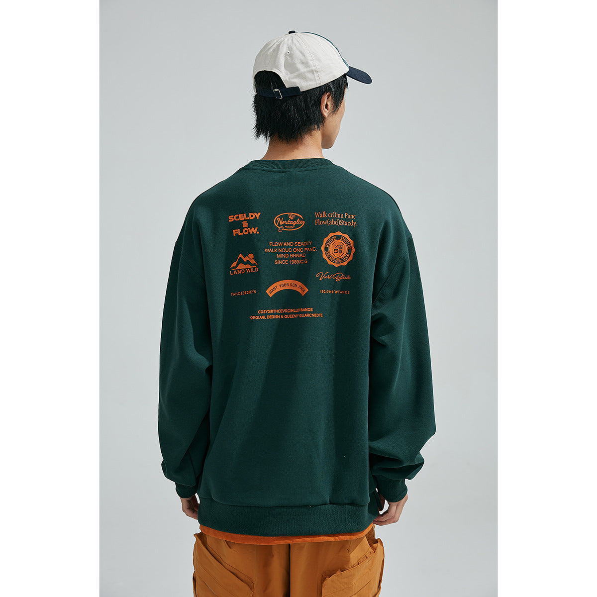 Back view of a model wearing a dark green sweatshirt with multiple orange graphic prints and ochre shorts.