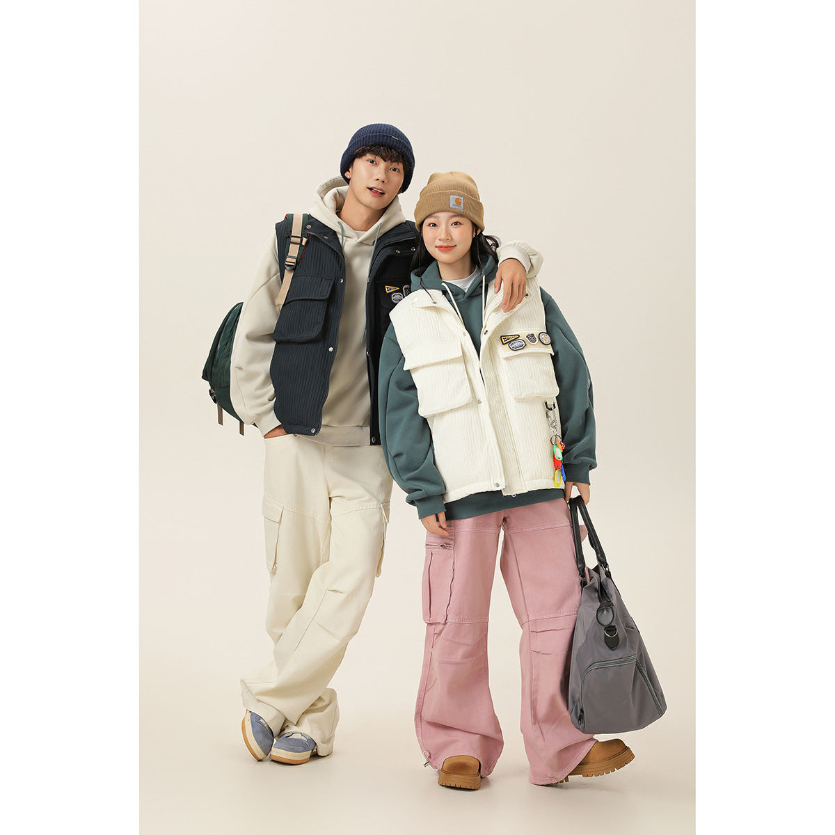 Two models wear utility vests: a navy vest over a cream hoodie and an ivory vest over a teal hoodie.