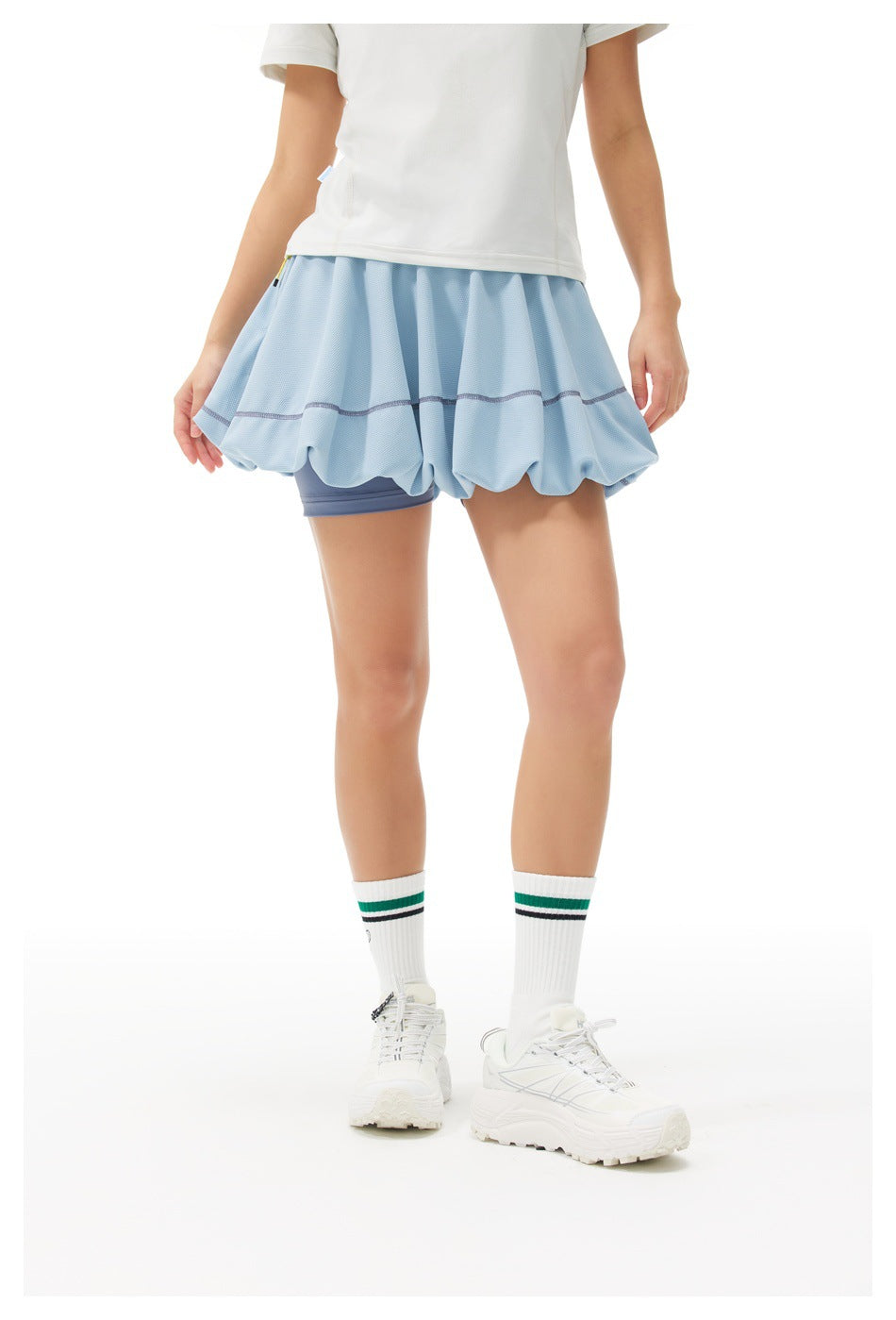 Front view of a model wearing a light blue bubble tennis skirt with dark blue built-in shorts and a white top.