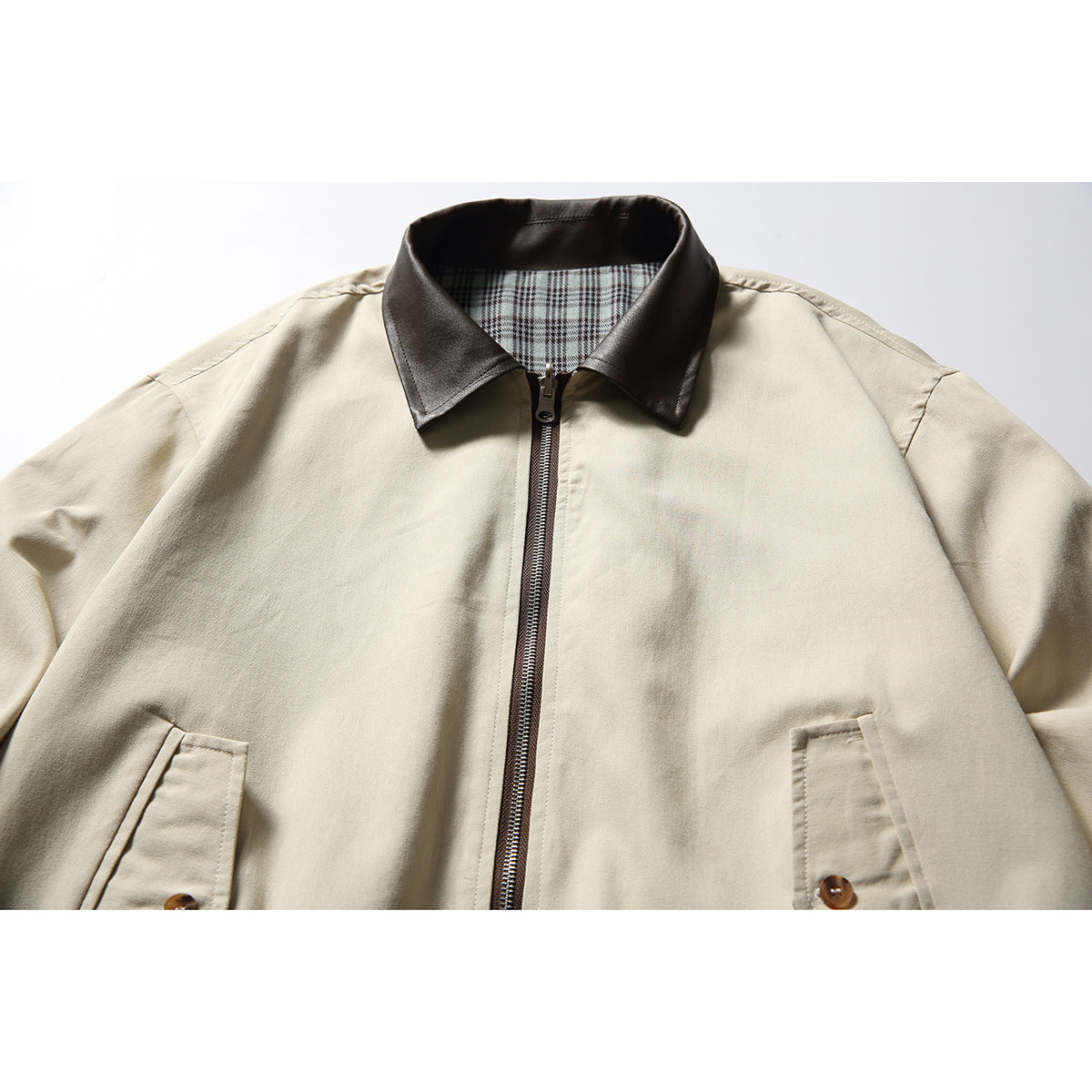 Close-up of a light beige jacket with a dark brown collar, plaid inner lining, front zipper and angled pockets.