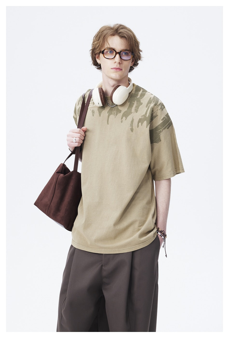 Model wearing khaki green T-shirt with distressed shoulder print, headphones around neck and brown tote bag, upper-body view
