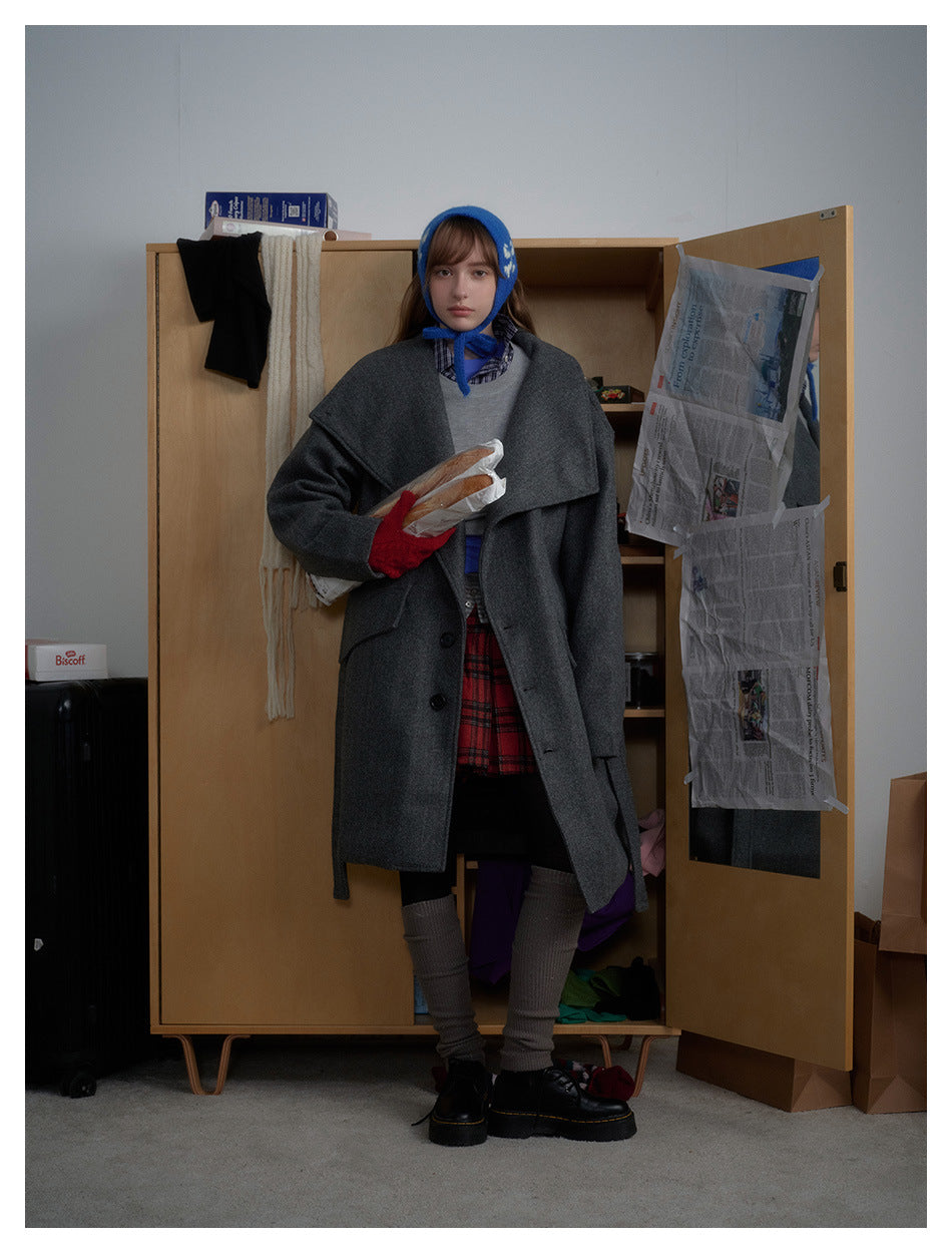 Model wearing the gray belted coat open over a gray sweater, plaid skirt and boots, standing by a wooden wardrobe