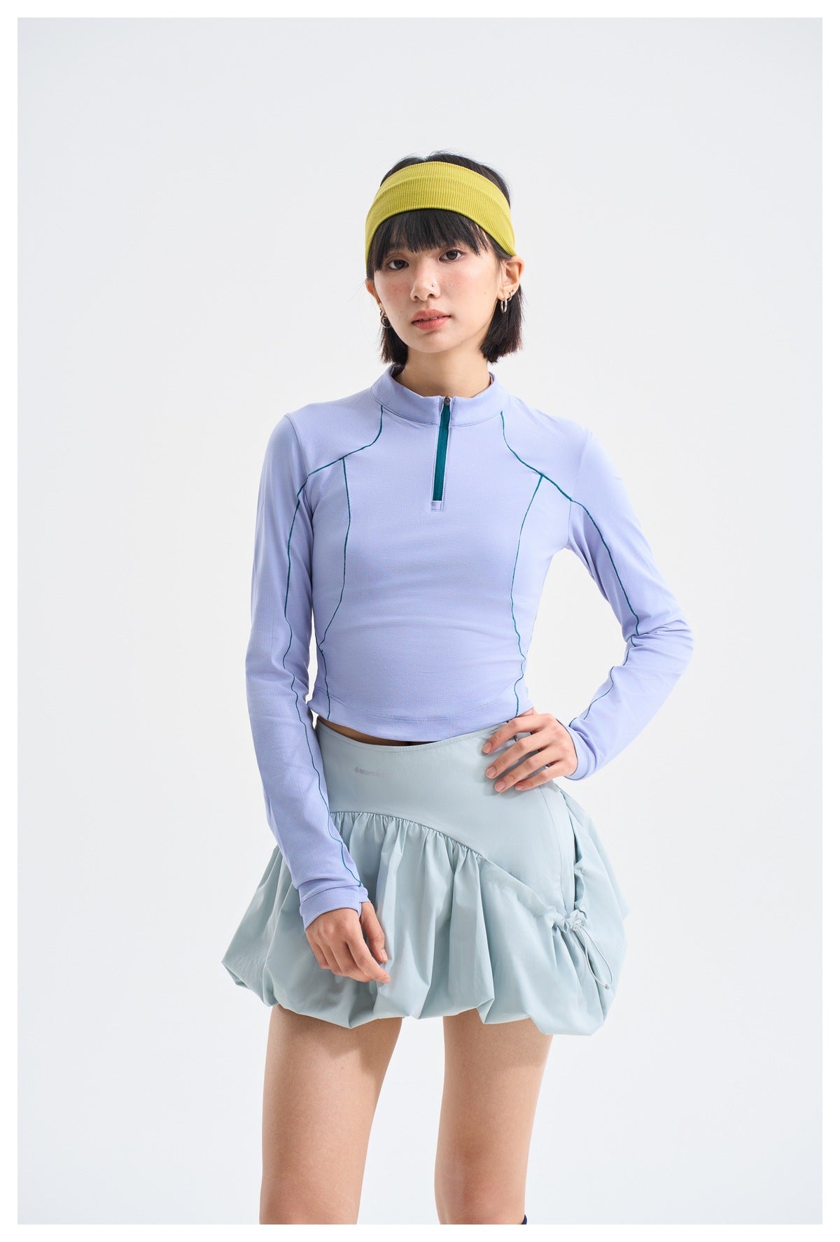 Model wearing a lilac long-sleeve quarter-zip top with teal seam piping and a pale blue gathered skirt.