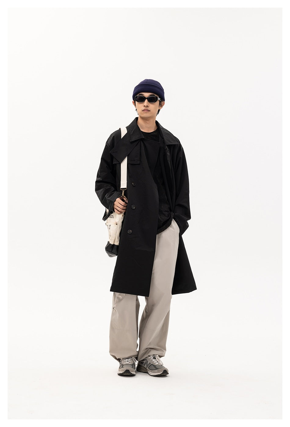 Model wearing an open black long trench coat over a black tee with light gray pants, carrying a canvas bag