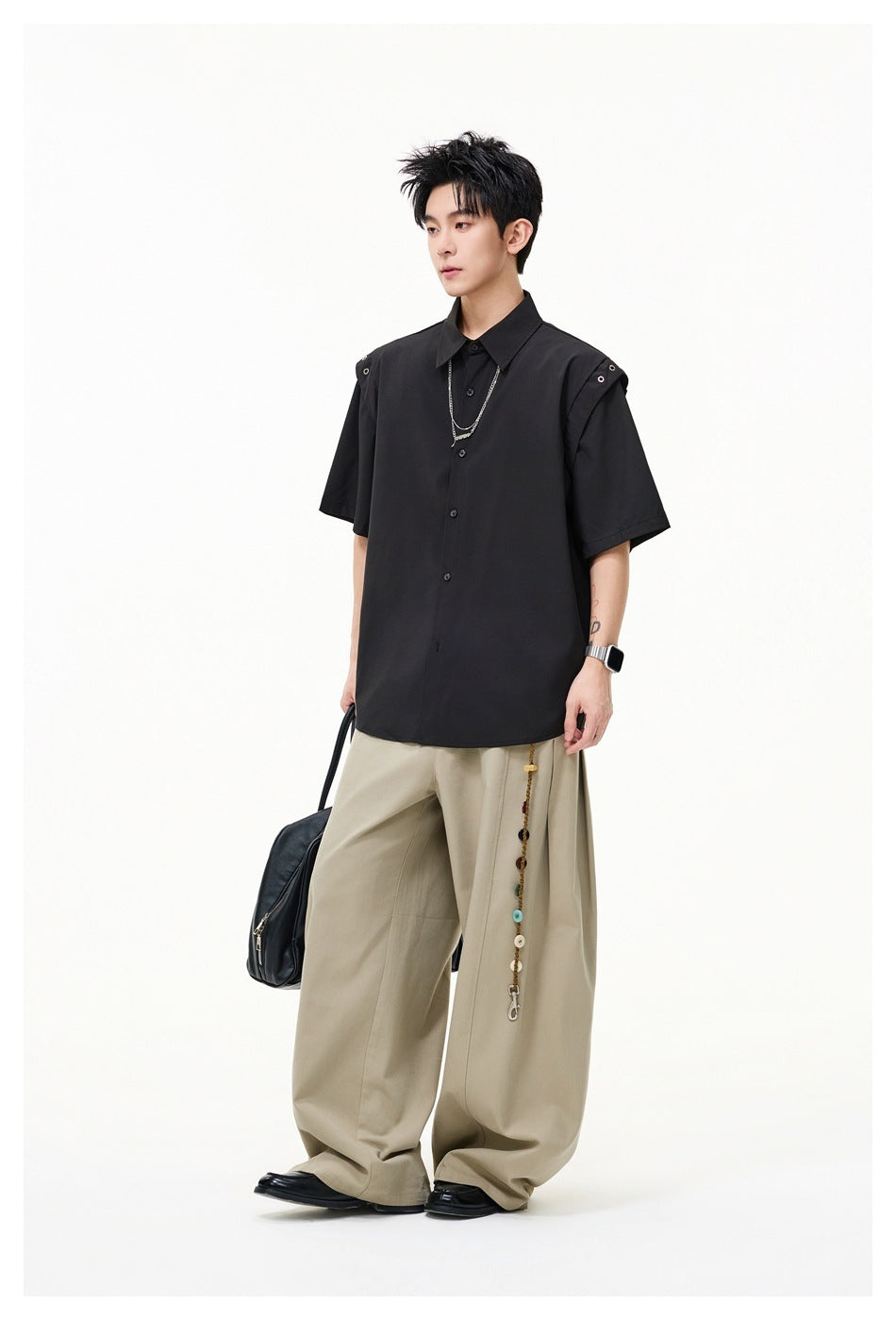 Full-length view of model wearing a black short-sleeve button-up shirt with metal-eyelet shoulder tabs and beige wide-leg