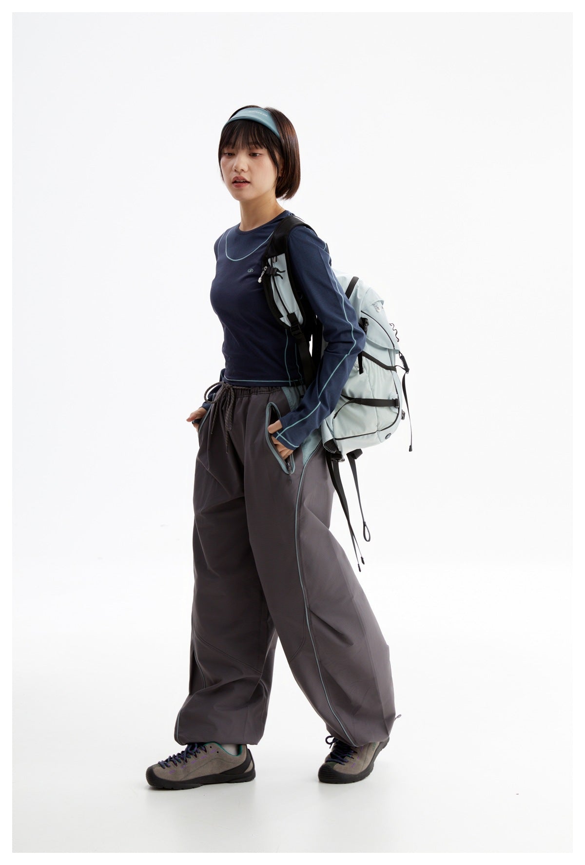 Model wearing a navy long-sleeve top with teal piping and charcoal wide-leg pants, carrying a pale blue backpack.