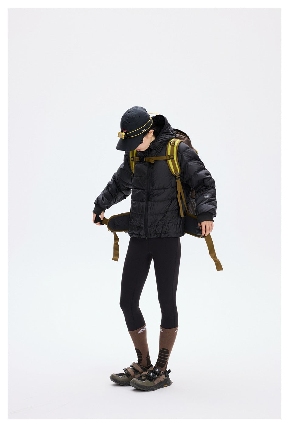 A model in a black hooded puffer jacket and hiking backpack adjusts the backpack's waist belt.
