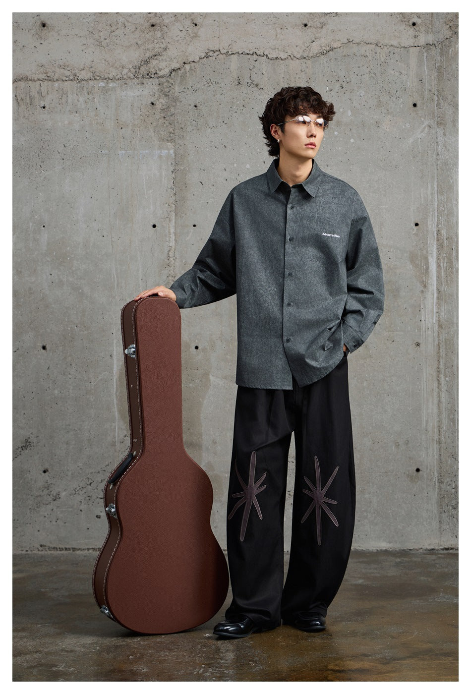 Model wearing a charcoal button-up shirt with small white chest lettering and black wide-leg trousers with starburst designs.
