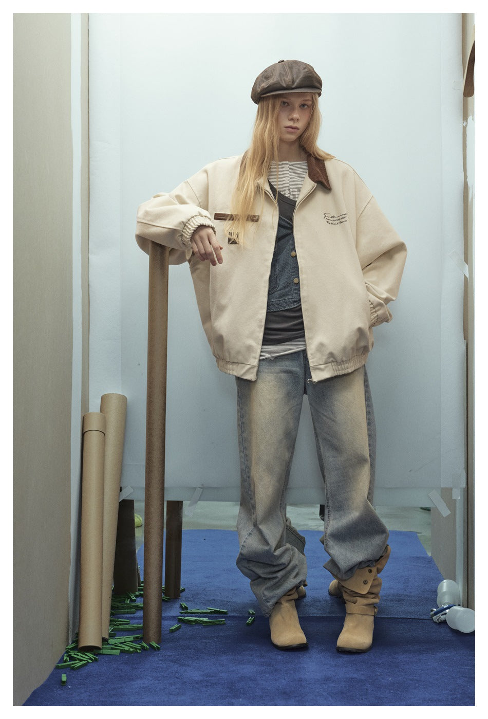 Model standing in an oversized light khaki jacket with brown collar over layered tops and wide faded jeans