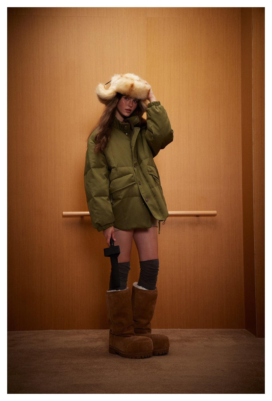 Full-length model wearing an olive-green padded button-front jacket, cream fur hat, dark leg warmers, and brown boots.
