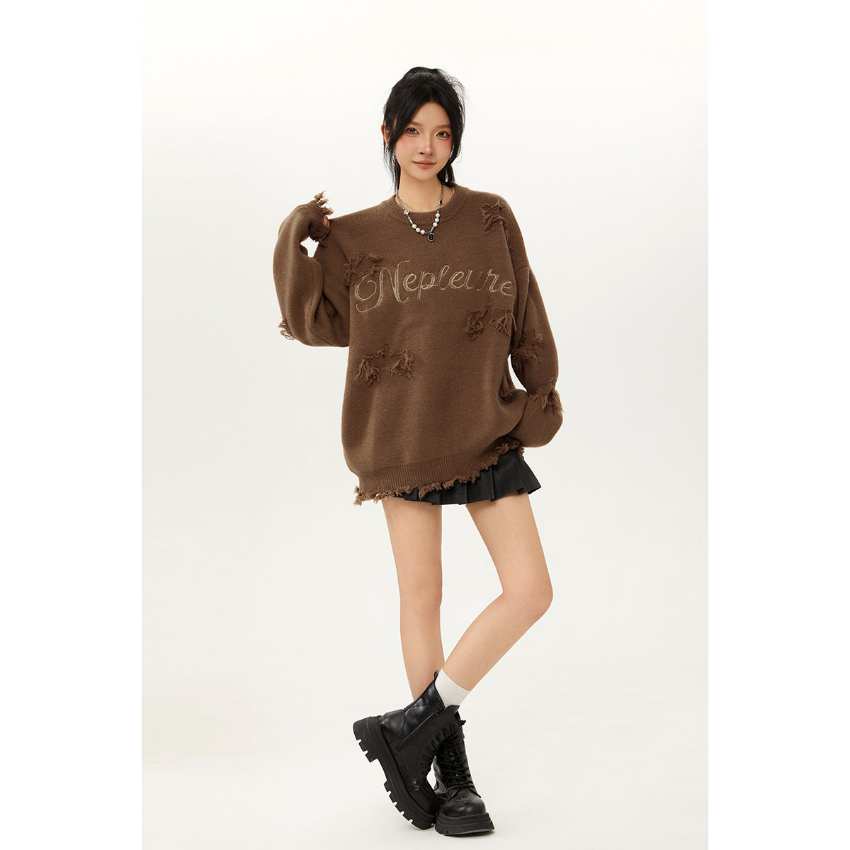 Model wearing an oversized brown sweater with distressed patches and script lettering, paired with a black skirt and boots.