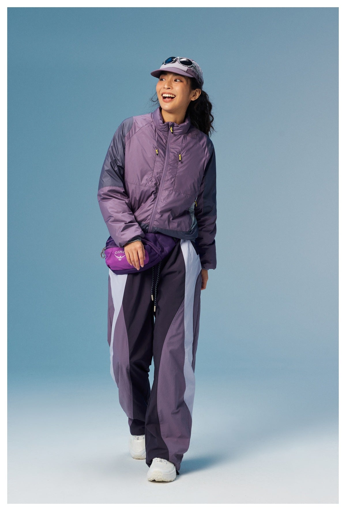 Model wearing a lavender color-block zip jacket and matching wide-leg pants, with a cap and waist bag.