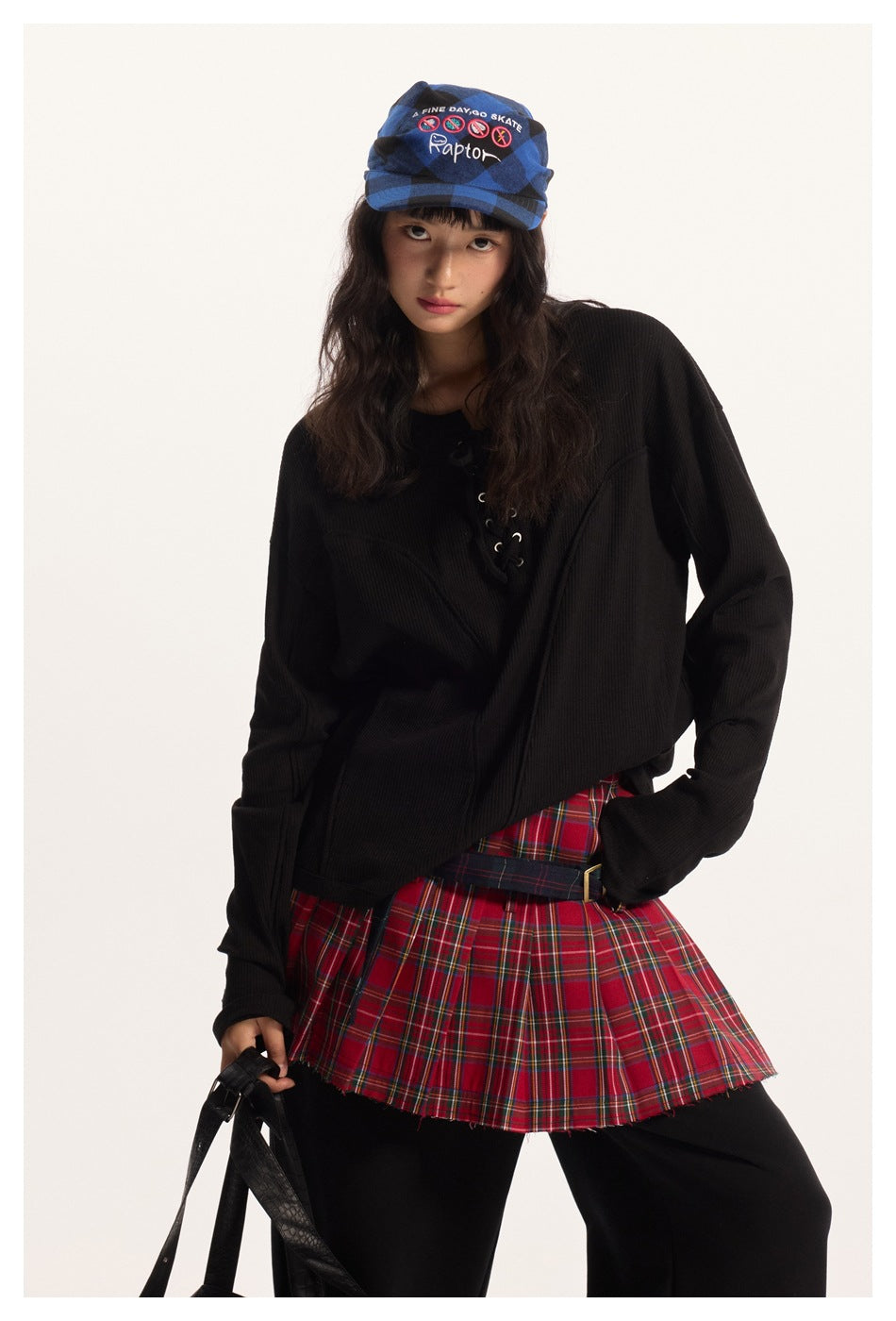 Model wearing an oversized black ribbed lace-up top with a red plaid pleated skirt and blue patterned cap.
