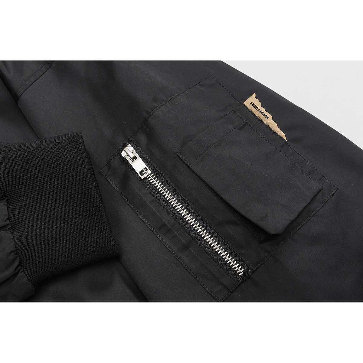 Close-up of a black jacket sleeve pocket with a flap, silver zipper, and small tan fabric label.