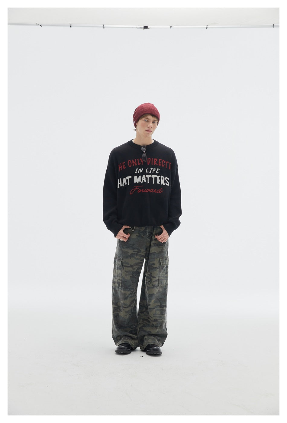 Full-length model in a black lettered crewneck sweater, burgundy beanie, and loose camouflage cargo pants.