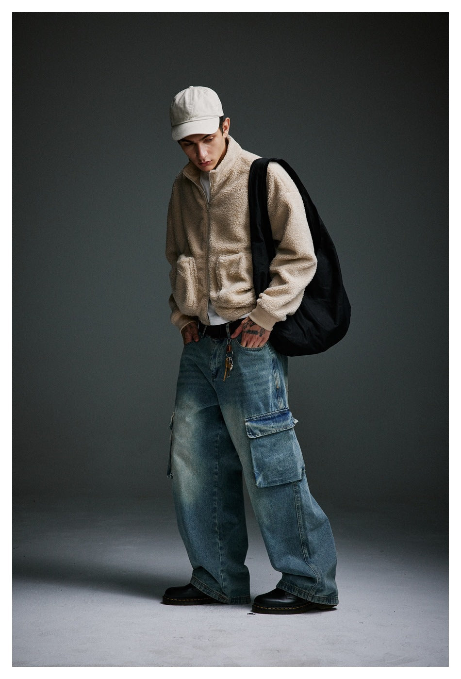 Model wearing a cream zip-up jacket, oversized blue cargo jeans, white cap and black shoulder bag.