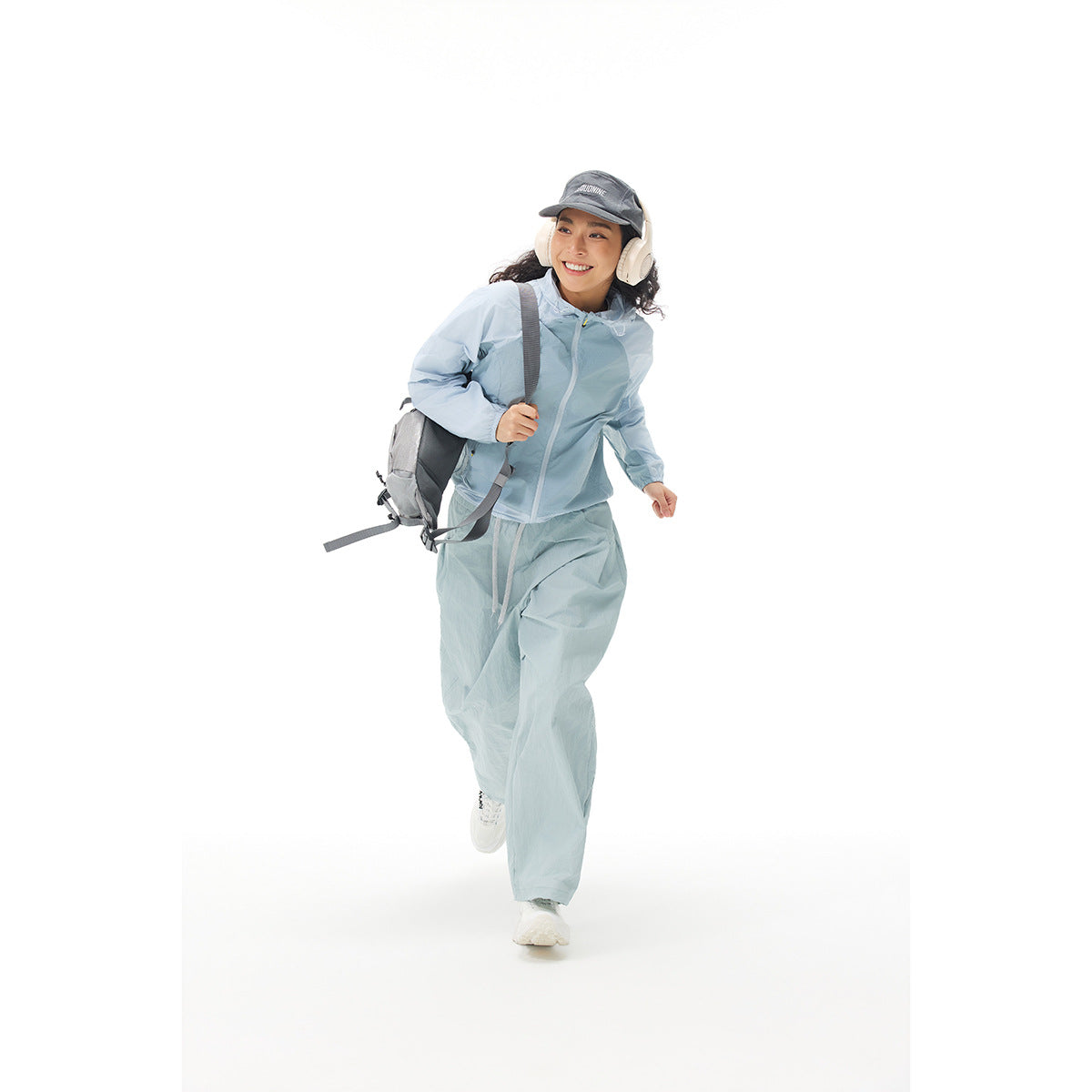 Woman running in a pale blue hooded jacket and matching loose pants, with a backpack, cap and headphones.