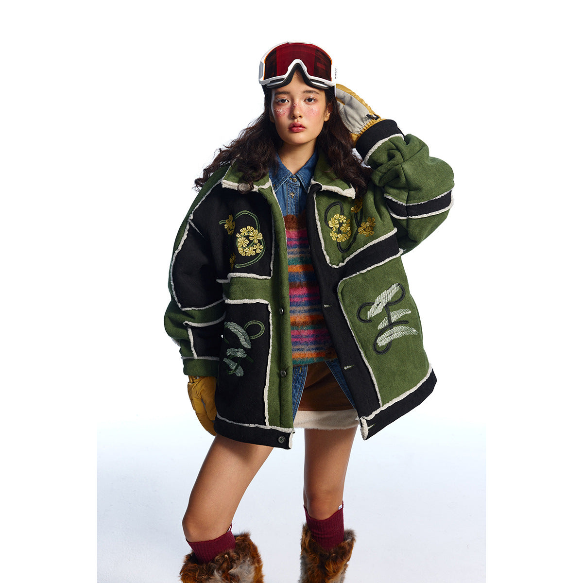Model wearing an oversized green and black jacket with cream trim and floral embroidery, styled with ski goggles.