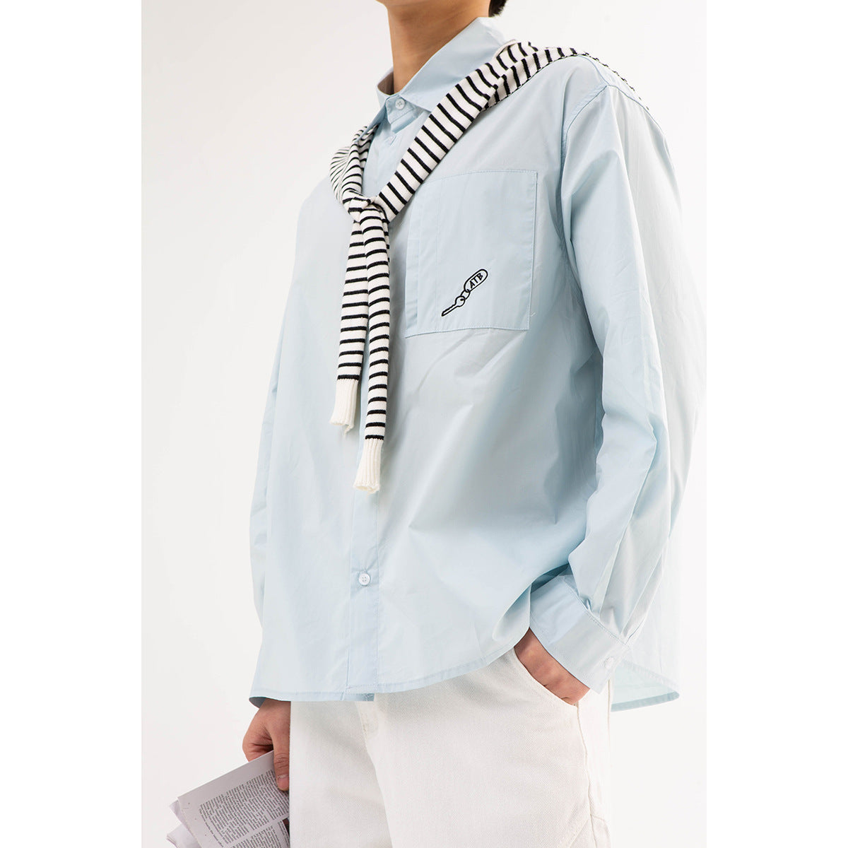 Cropped model wearing a pale blue button-up shirt with a chest pocket and a black-and-white striped sweater draped over the