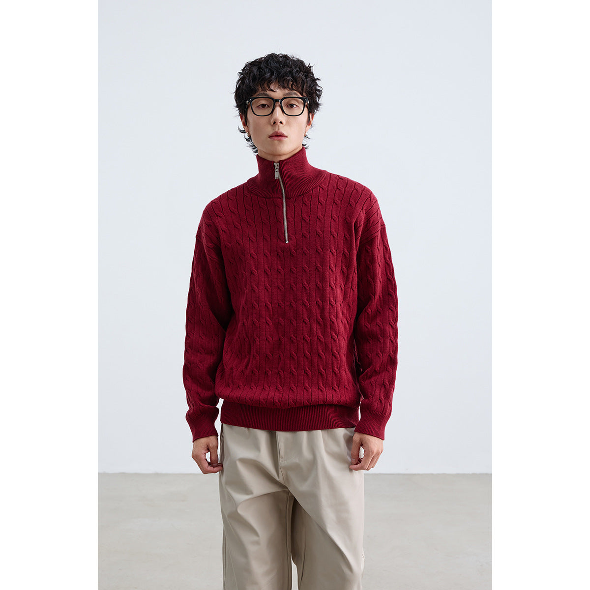 Model wearing a burgundy cable-knit quarter-zip sweater with a high collar and beige trousers.