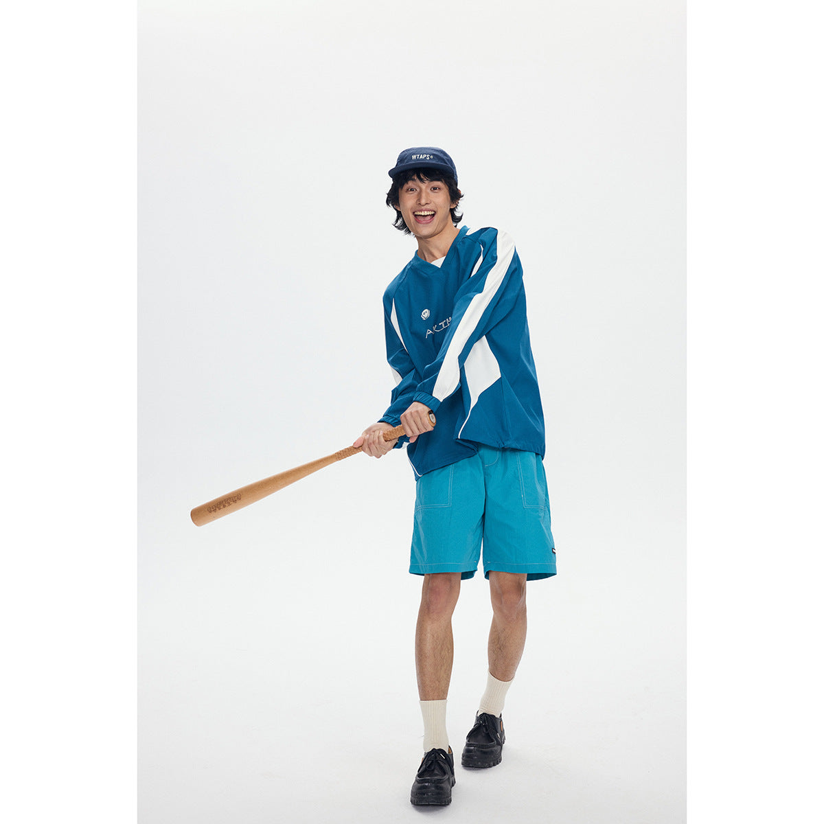 Model wearing a teal V-neck pullover with white sleeve panels and matching teal shorts, holding a baseball bat.