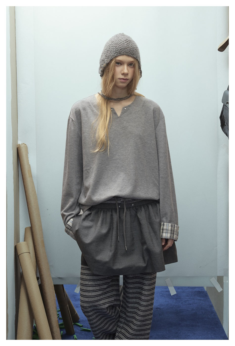Front view of a model wearing a loose gray notched-neck top with plaid cuffs, dark shorts, striped trousers, and a gray knit