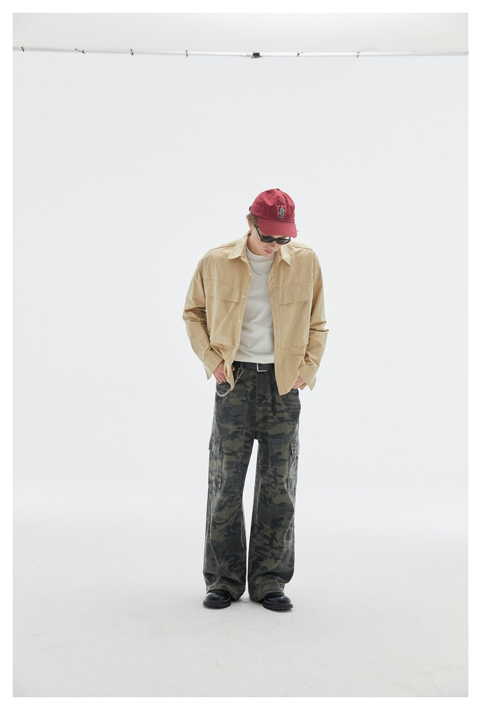 Model looking down in a beige button-up overshirt and loose dark camouflage cargo pants.