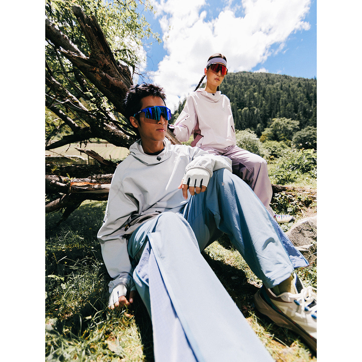 Two models outdoors in pale technical tops and loose pants; the seated model wears blue-gray, and the other wears pink.