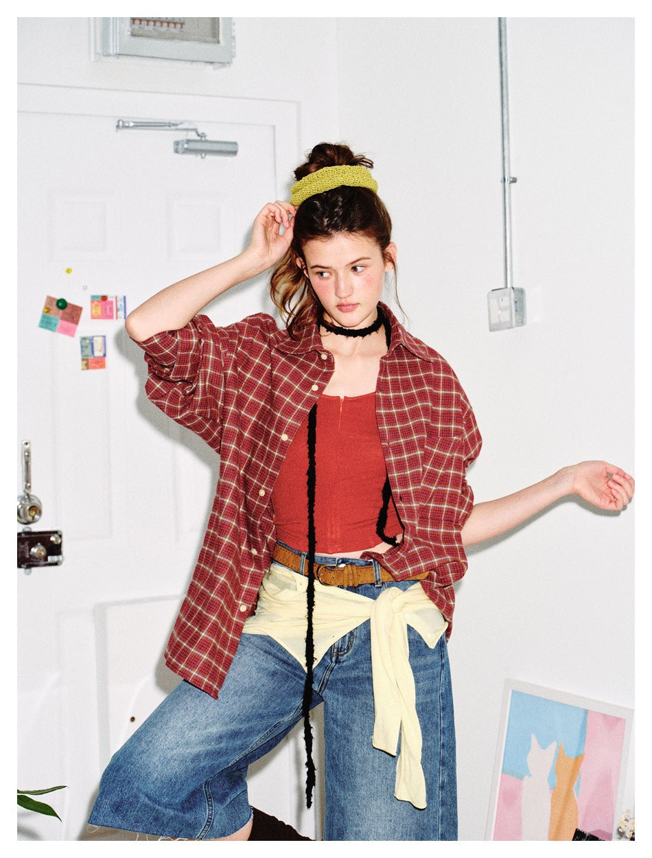 Model wearing an open burgundy plaid shirt over a red top and wide-leg denim shorts, hand raised to hair
