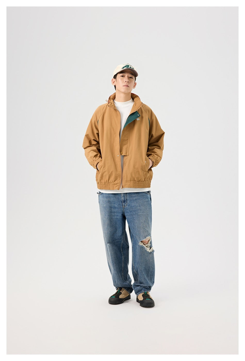 Model wearing the tan color-block zip jacket open over a white tee with blue ripped jeans, standing front view