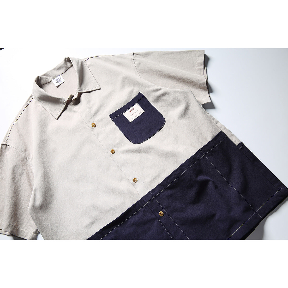 Angled flat lay of a short-sleeve button-up shirt with a light gray upper half, navy lower panel, and navy chest pocket.