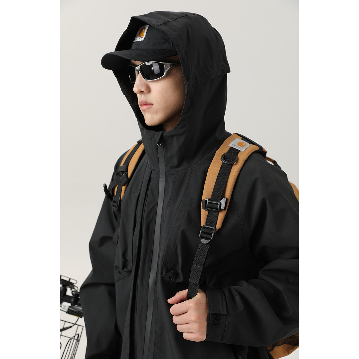 Model wearing a black hooded jacket with the hood over a cap, sunglasses, and a backpack.