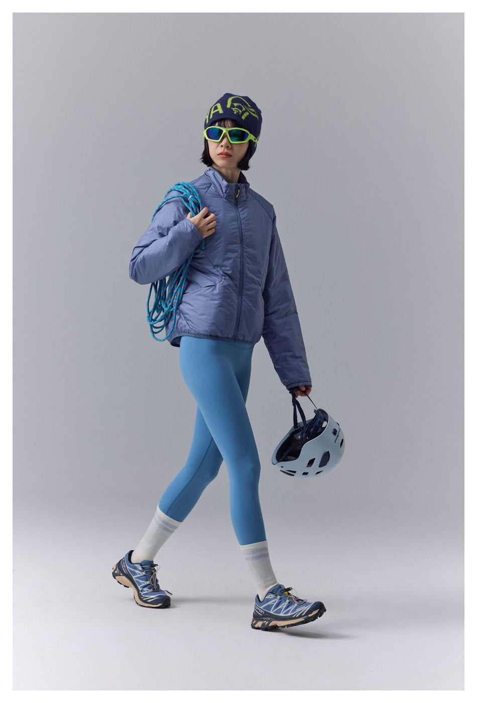 Model walking in a periwinkle-blue padded jacket and blue leggings, carrying a climbing rope and helmet.