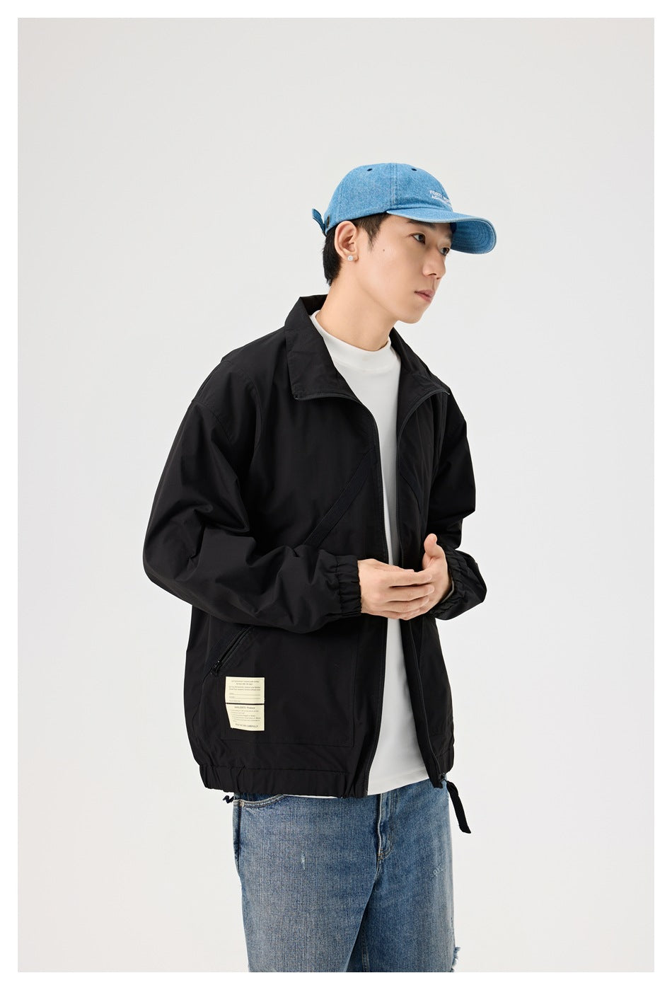 Three-quarter view of a model wearing an open black jacket over a white shirt, with blue jeans and a blue cap.