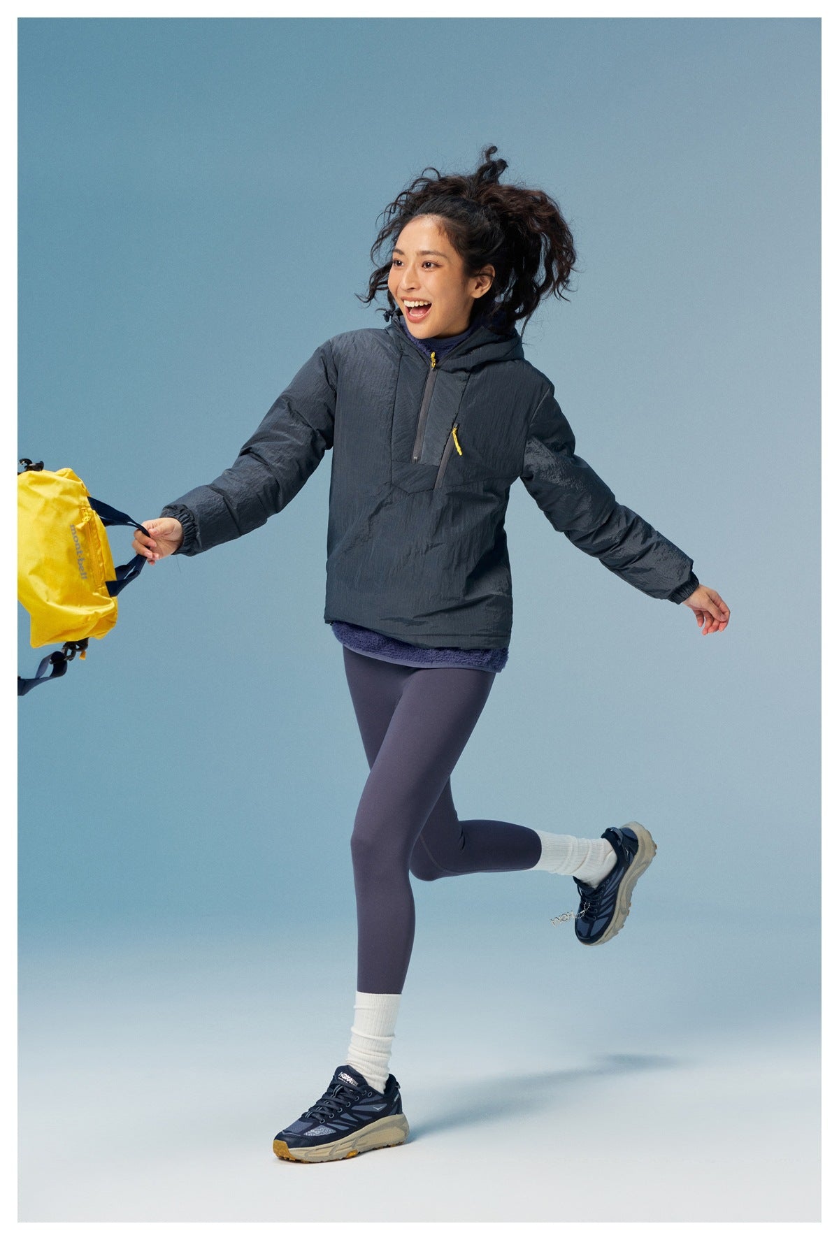 Woman wearing a charcoal hooded pullover with yellow zipper pulls and purple leggings, posed against a blue studio