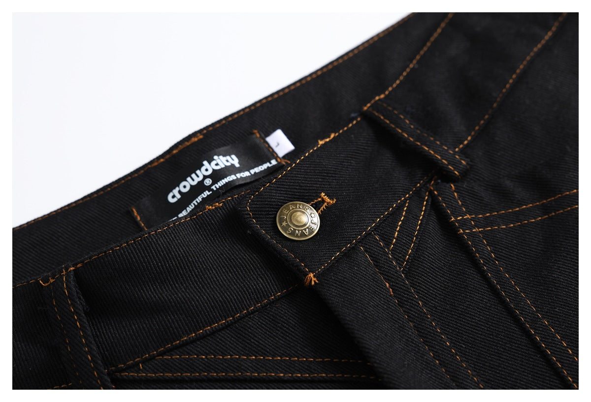 Close-up of black jeans showing the waistband label, metal button, fly, and orange stitching.
