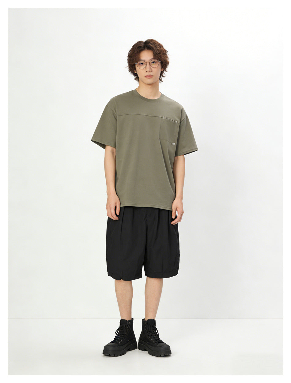 Model wearing an olive short-sleeve pocket T-shirt with black shorts and black high-top boots.
