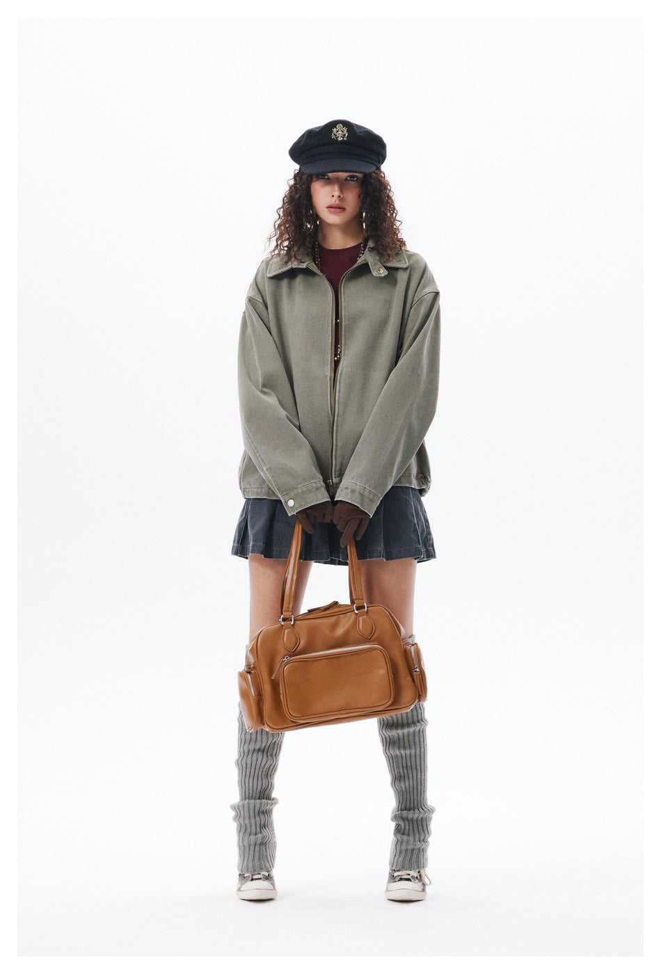 Model wearing the green zip jacket with a dark cap, pleated denim mini skirt, gray leg warmers and a tan bag
