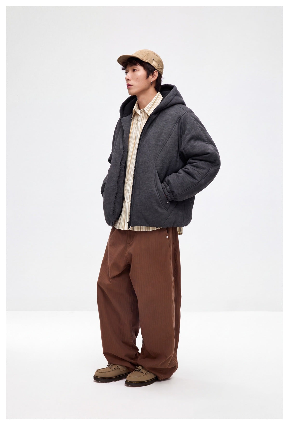 Model wearing a charcoal hooded zip jacket with curved front seams and pockets, styled with brown trousers.