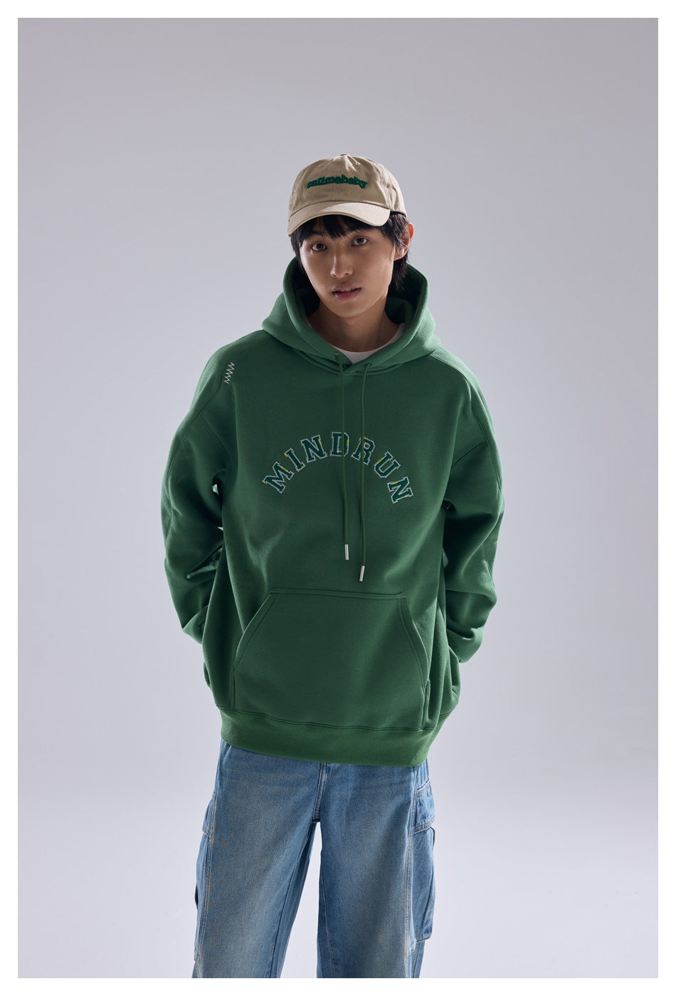 Model facing forward in a green MINDRUN hoodie with a beige cap and light-wash cargo jeans