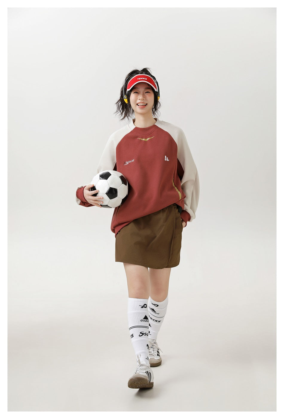Smiling model holding a soccer ball wearing a brick red raglan sweatshirt with cream sleeves and a brown skirt