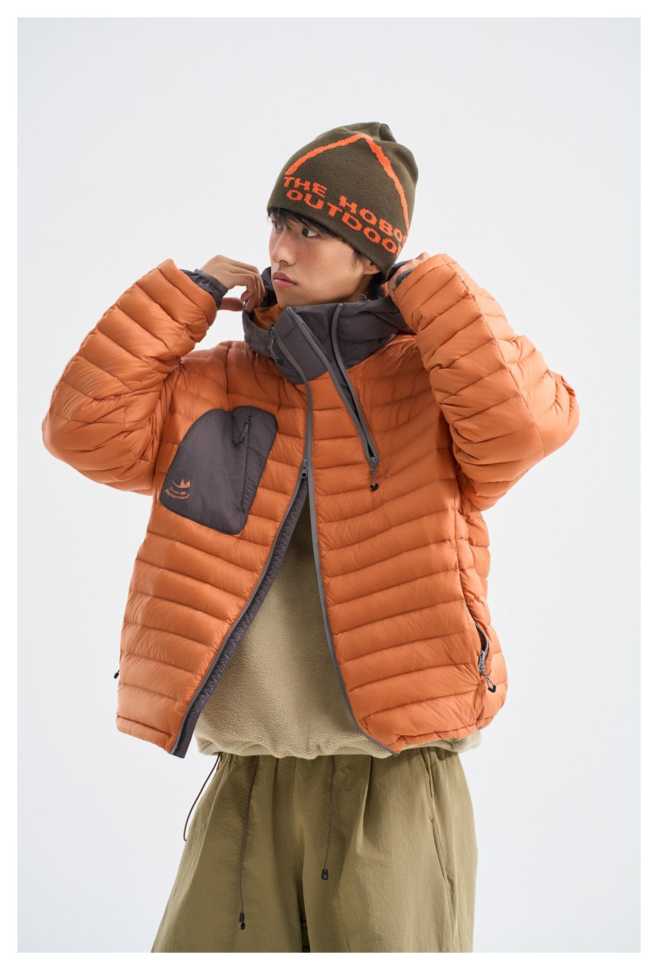 Model wearing an orange quilted puffer jacket with a gray hood and olive pants, adjusting the collar.