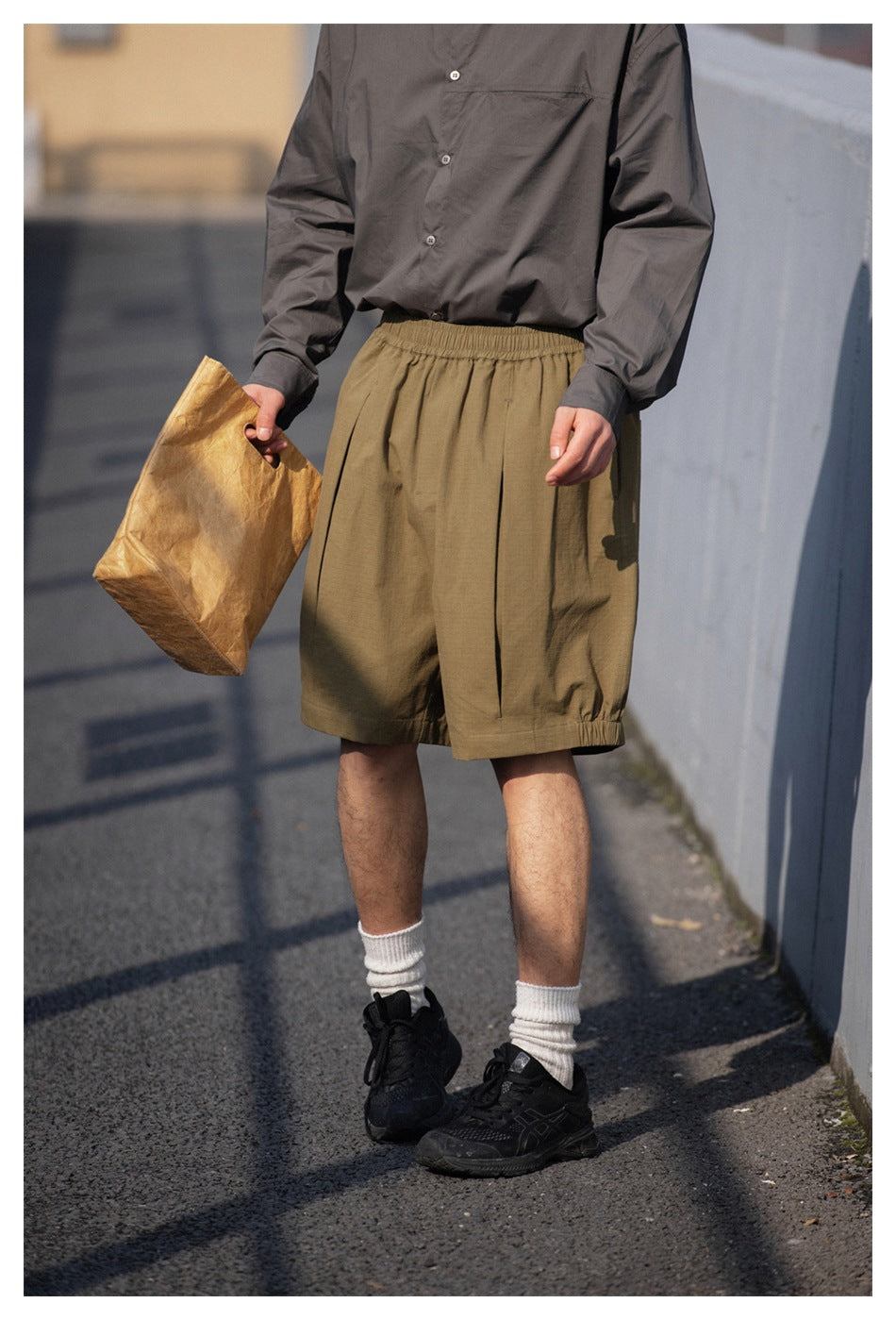 Person in olive pleated knee-length shorts and gray button shirt holding a paper bag outdoors, torso-to-feet crop