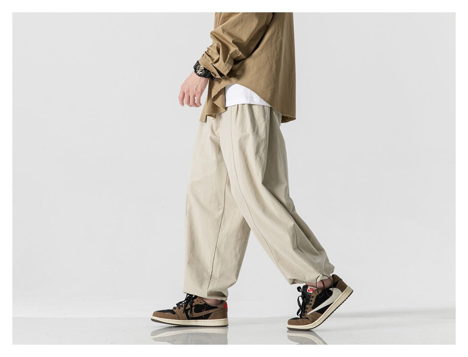 Cropped side view of a model wearing loose light beige trousers with an elastic waist and gathered hems.