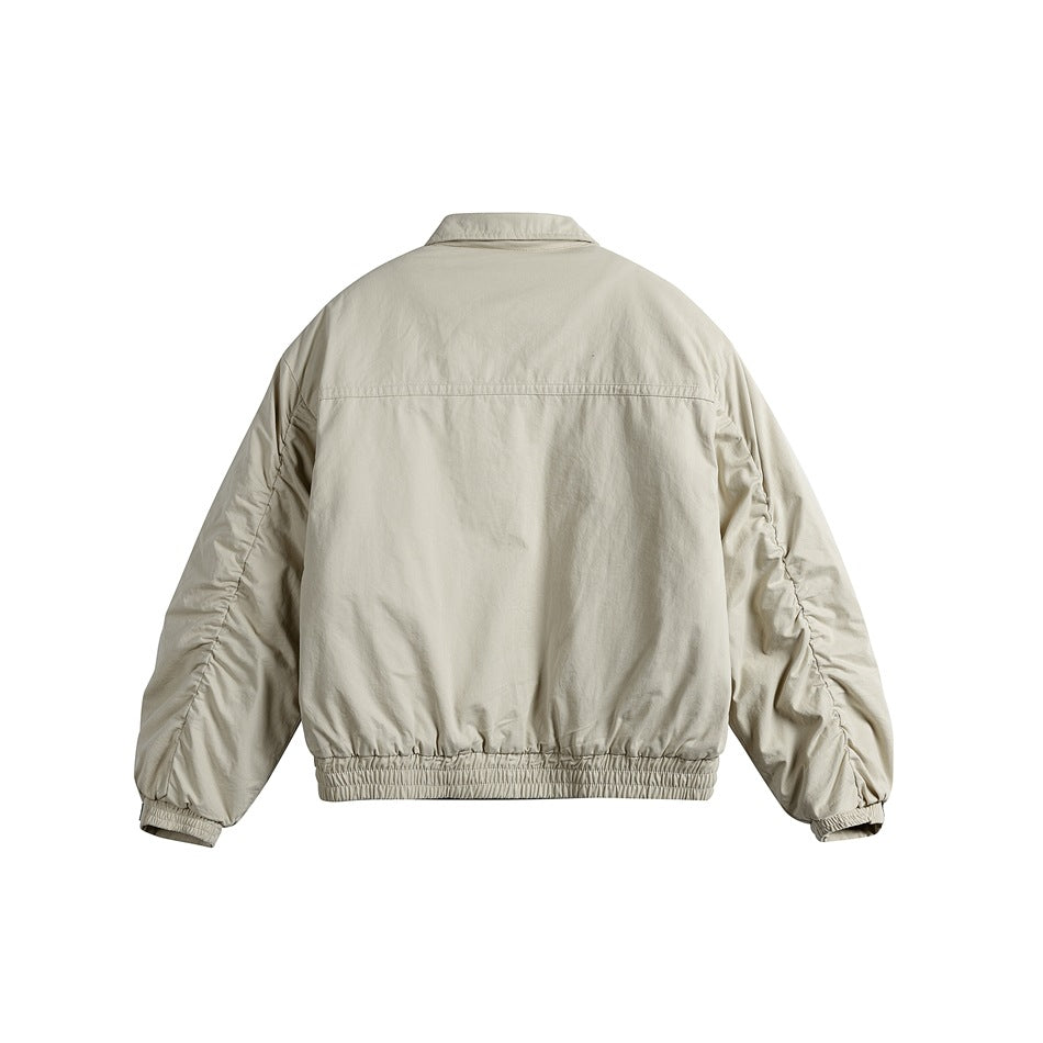Back view of a light beige jacket with a shoulder seam, gathered cuffs, and elastic hem.