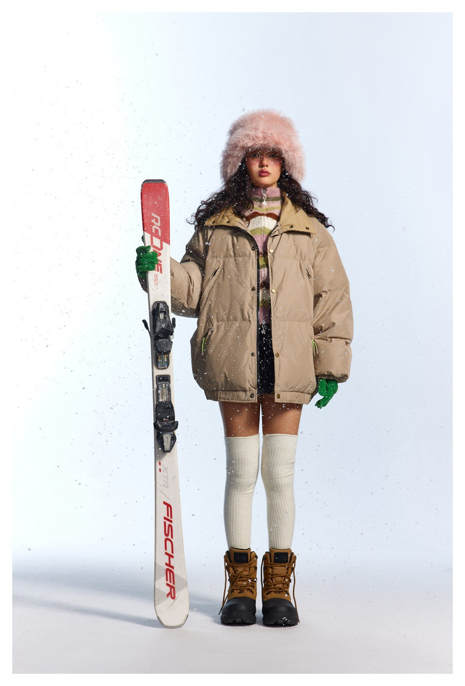 Full-length model wearing a light beige puffer jacket with a high collar, posed with a ski against a snowy white backdrop.