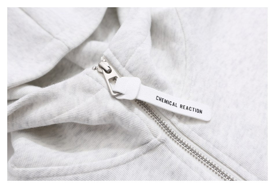 Close-up of a light gray heathered garment zipper with a white pull tab printed CHEMICAL REACTION.