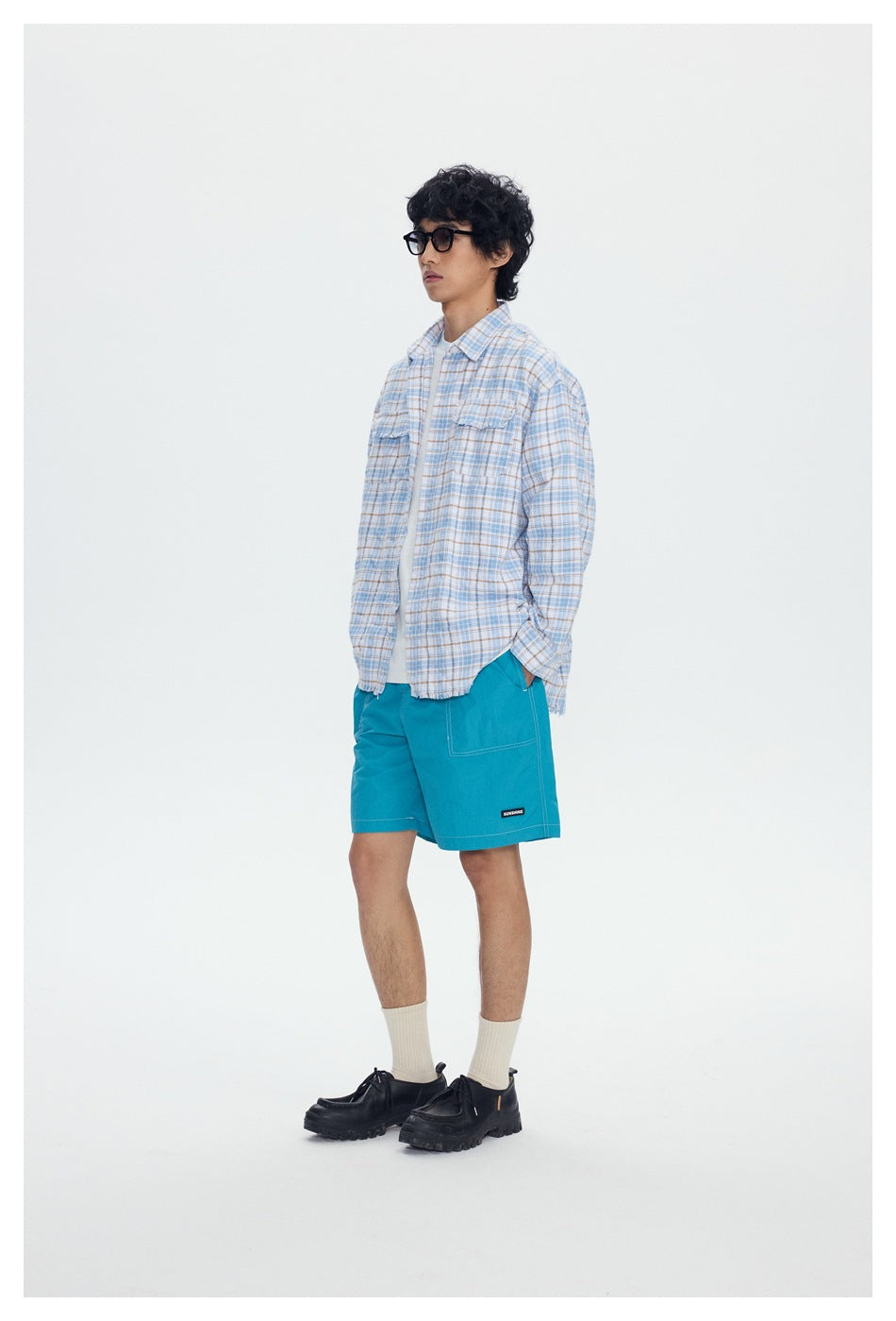 Model wearing an open blue plaid shirt over a white tee with teal shorts, side view